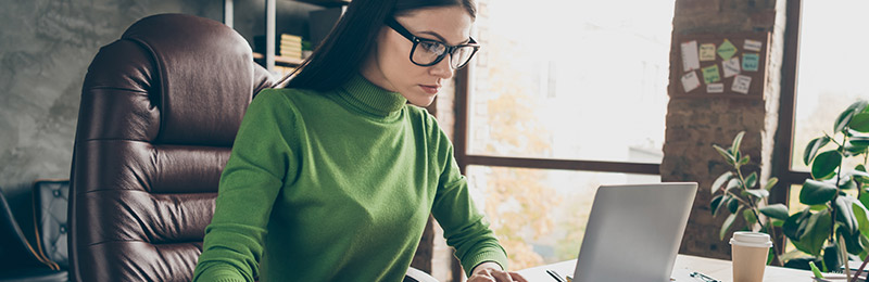 Female entrepreneur sitting at her desk next to a window. Trying to write her business plan in under an hour using a laptop.
